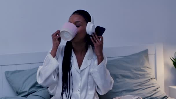 Wanita Afrika Amerika cantik menikmati mendengarkan musik dengan headphone putih besar menggunakan smartphone dan minuman dari cangkir. Wanita hitam berpose di kamar tidur dengan piyama. Tutup. Gerakan lambat. — Stok Video