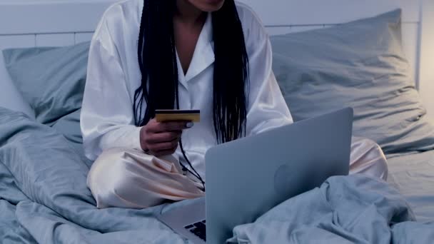 Mujer bastante afroamericana hace la compra en línea utilizando un portátil y una tarjeta de crédito. Mujer negra posando en el dormitorio en la cama en pijama. Luz interior de la habitación del hogar. De cerca. Movimiento lento. — Vídeo de stock