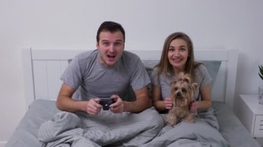 Mutlu genç çift küçük köpekle yatakta yatıyor. Tutkulu bir adam kablosuz kontrolörle oyun oynar ve kazanır. Parlak yatak odasında poz veren sevgili portresi. Aile eğlencesi. Yavaş çekim