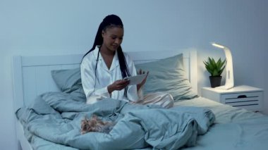 Afro-Amerikalı kadın içeriği dijital tabletten görüyor, mesaj yazıyor, sosyal ağda iletişim kuruyor. Siyah kadın, yatakta pijamalarıyla köpeği Yorkshire Teriyeri 'yle poz veriyor. Yavaş çekim