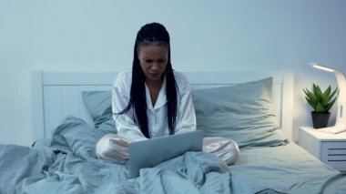 Güzel bir Afrikalı Amerikalı kadın dizüstü bilgisayardan içeriği görüyor, bir mesaj yazıyor, sosyal ağda iletişim kuruyor. Siyah kadın pijamalarıyla yatak odasında poz veriyor. Yavaş çekim.