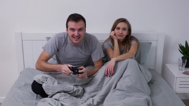 Portret młodej szczęśliwej pary pozującej leżącej w sypialni na łóżku. Mężczyzna z pasją gra z kontrolerem bezprzewodowym i przegrywa, kobieta jest znudzona i pociesza przegranego. Zwolniony ruch. — Wideo stockowe