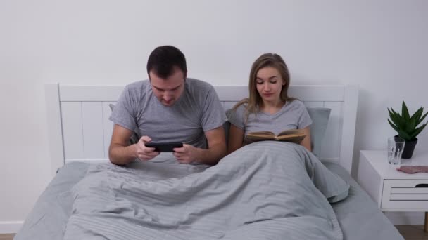 Retrato de casal jovem feliz posando enquanto deitado no quarto na cama. Homem com paixão joga jogo no celular e ganha com prazer wow, mulher lê livro e, em seguida, deliciosamente se alegra. Movimento lento. — Vídeo de Stock