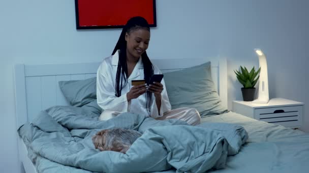Mulher muito afro-americana faz uma compra on-line usando um telefone celular e um cartão de crédito. Mulher negra posando no quarto na cama de pijama com seu cachorro Yorkshire Terrier. Movimento lento. — Vídeo de Stock