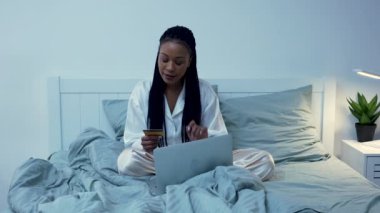 Güzel bir Afrikalı Amerikalı kadın taşınabilir bir laptop ve kredi kartı kullanarak online alışveriş yapıyor. Siyah kadın pijamalarıyla yatak odasında poz veriyor. Odanın içi aydınlık. Yavaş çekim.