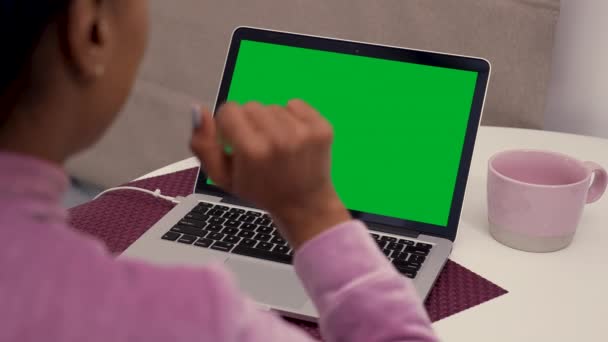 Över axeln skott av svart kvinna sitter vid bordet och gör videosamtal på laptop med grön chroma key screen. Ung kvinna vinkar hej och hej kommunicerar online via webbkamera. Närbild. Långsamma rörelser. — Stockvideo