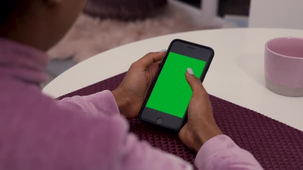 Jonge Afro-Amerikaanse vrouw zit aan tafel in de kamer en maakt gebruik van smartphone met groene scherm chroma sleutel. Weinig soorten beweging - scrollen, tikken, in- en uitzoomen. Sluit maar af. Langzame beweging. — Stockvideo