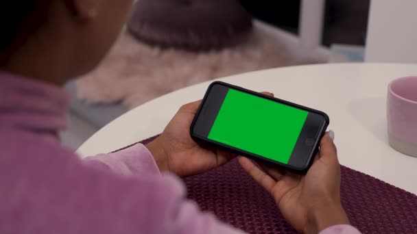 Jovem afro-americana senta-se à mesa no quarto e usa smartphone com chave chroma tela verde. Poucos tipos de movimento - rolagem, tocando, zoom in e out. Fecha. Movimento lento. — Vídeo de Stock