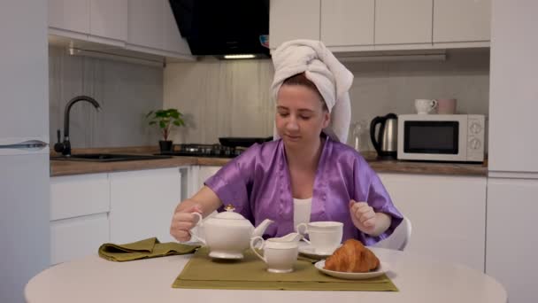 Jovem está sentada à mesa na cozinha, derrama-se xícara de café com creme, desfrutando de aroma e sabor da bebida. Feminino em toalha branca na cabeça posa contra o fundo da sala de luz. Movimento lento — Vídeo de Stock