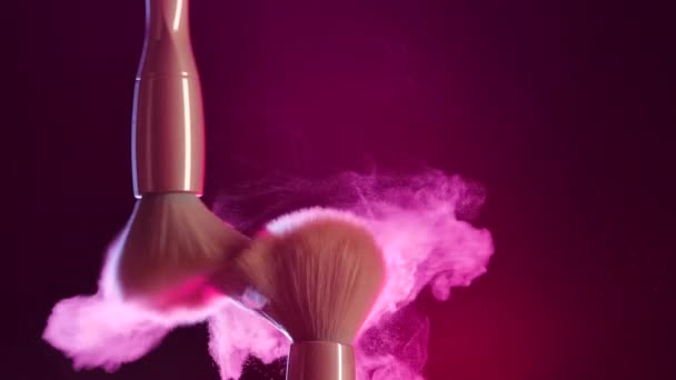 Dos pinceles de maquillaje que se tocan forman un hermoso remolino de partículas de polvo cosmético. Primer plano de pinceles de maquillaje en estudio oscuro en luz de neón rosa. Movimiento lento. — Vídeo de stock