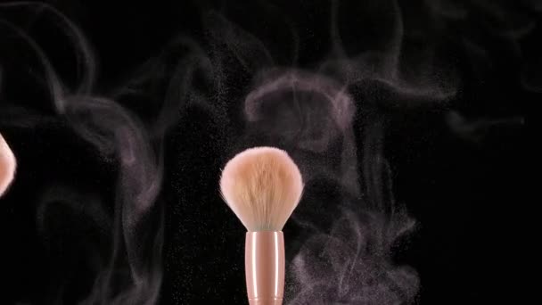 Duas escovas de maquiagem colidem e causam um turbilhão de partículas de pó cosmético contra um fundo preto. O pó fino do pó esmagado voa lindamente no ar. Fecha. Movimento lento. — Vídeo de Stock