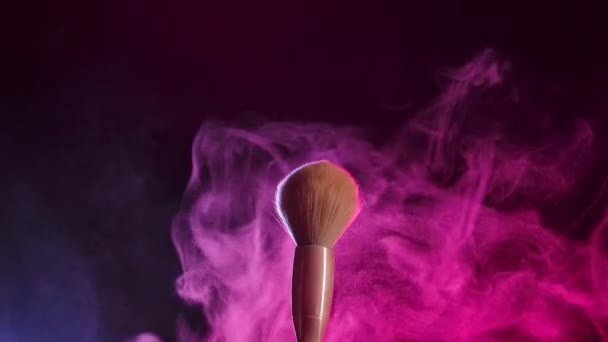 Zwei Make-up-Pinsel prallen aufeinander und verursachen einen Strudel aus Puderpartikeln vor schwarzem Hintergrund in rosa Neonlicht. Feinstaub des zerkleinerten Pulvers fliegt schön in der Luft. Nahaufnahme. Zeitlupe. — Stockvideo