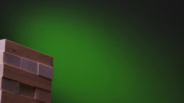 Yeşil gradyan üzerinde Jenga oyunu. Kızlar yavaş çekimde kuleye bir blok bildiriyor. Kamera sağdan sola hareket ediyor. Kapat..