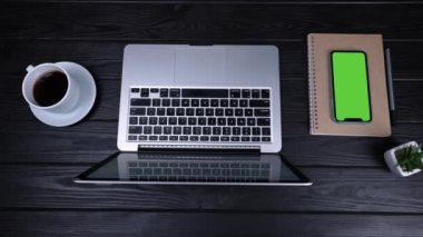 Masanın üzerinde açık bir dizüstü bilgisayar, bir fincan kahve, bir not defteri, bir kalem ve yeşil ekranlı bir akıllı telefon ve bir krom anahtar var. Çalışmak için hazırlanan işyerinin üst görüntüsü. Ağır çekim. Kapat..