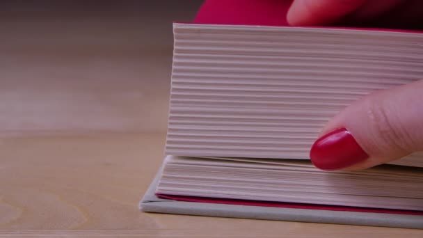 Een dik boek met rode titelpagina 's ligt op een houten tafel. Het meisje bladert er doorheen met haar duim. Langzame beweging. Sluiten.. — Stockvideo