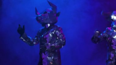 Aynalardan parlayan gümüş kostümlü dansçılar dumanlı stüdyo arka planında boynuzlu boğa şeklinde. Dans eden aktörler ve neon mavisi pembe ışıkta neşeyle sergilenen gösteriler. Kapatın. Yavaş çekim.