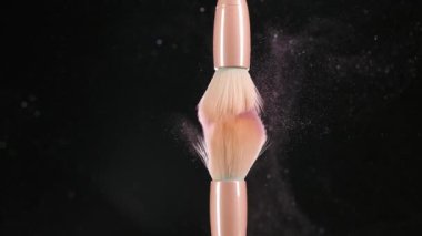 Two makeup brushes with pink powder, blush on a black background. In slow motion, the brushes touch each other, and small particles of makeup fly in the air, forming a swirl of sparkles. Close up.