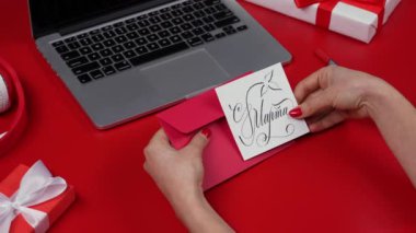 Kadın eli, içinde 8 Mart 'ın kutlaması olan bir kartpostalı zarfa koydu. Dizüstü bilgisayarı ve hediye kutuları olan kırmızı bir masa. Uluslararası Kadınlar Günü konsepti. Kapatın. Yavaş çekim.