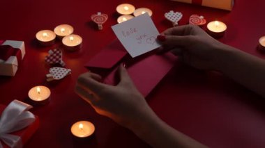 Womans hands put a valentines love postcard with the inscription love you in envelope. Top view of a red table with burning candles, decorative hearts and gift boxes. Close up. Slow motion.