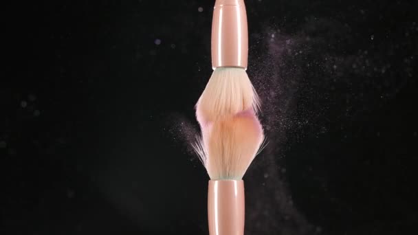 Two makeup brushes with pink powder, blush on a black background. In slow motion, the brushes touch each other, and small particles of makeup fly in the air, forming a swirl of sparkles. Close up. — Video