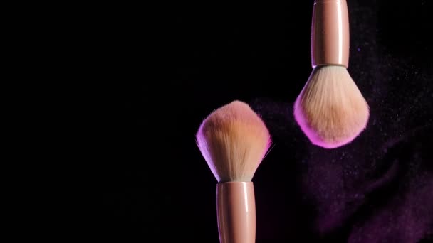 Desde el toque de dos pinceles de maquillaje, polvo brillante colorido o partículas de rubor flotan en el aire. Maquillaje creativo brillante, concepto sobre fondo negro en luz de neón rosa. En cámara lenta. De cerca.. — Vídeos de Stock