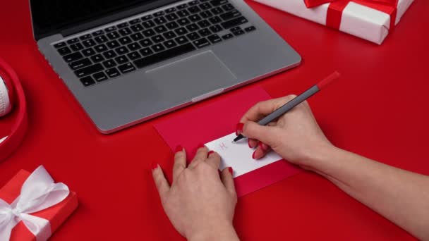 Las manos de las hembras escriben Feliz Día de la Mujer en marcador rojo en la postal de felicitación. Vista superior de una mesa roja con un portátil y cajas de regalo. Concepto del Día Internacional de la Mujer. De cerca. Movimiento lento. — Vídeo de stock