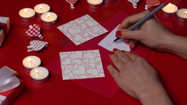 Vrouwenhanden schrijven Love you op een valentijnskaart. Bovenaanzicht van een rode tafel met brandende kaarsen, decoratieve hartjes en geschenkdozen. Romantische stemming. Valentijnsdag concept. Sluit maar af. Langzame beweging. — Stockvideo