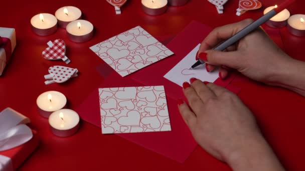 Womens hands draw a heart sign on a love valentine card. Top view of a red table with burning candles, decorative hearts and gift boxes. Valentines Day concept. Romantic mood. Close up. Slow motion. — Stockvideo