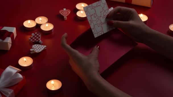 女性の手はお祝いの封筒にポストカードを愛するバレンタインを置く。燃えるろうそく、装飾的な心と贈り物と赤いテーブルの上のビュー。ライトとロマンチックな夕暮れの背景。閉めろ。スローモーション — ストック動画
