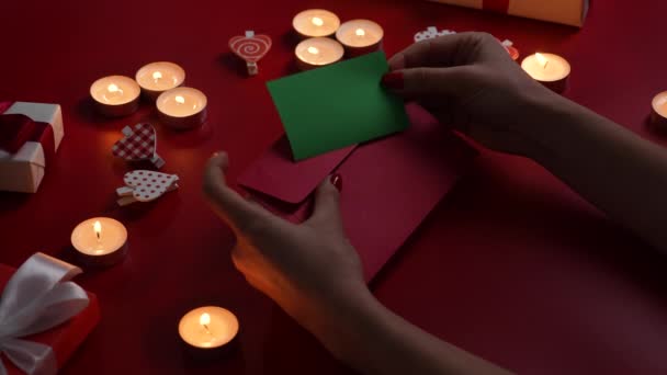 Womans hands put valentines postcard with green screen chroma key in envelope. Top view of red table with burning candles, decorative hearts and gift boxes. Romantic twilight. Close up. Slow motion. — Vídeo de stock