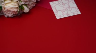 Beyaz kurdeleli bir hediye kutusu kırmızı masaya düşer. Sevgililer Günü 'nün özelliklerine sahip şenlikli bir geçmiş, bir buket gül, bir zarf, sevgililer günü ve dekoratif kalpler. Kapatın. Yavaş çekim.