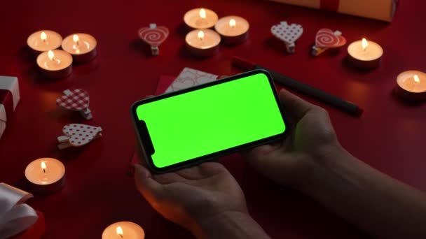 Womans hands hold smartphone with a green screen chroma key in horizontal position. Top view of a red table with burning candles, hearts and gift. Romantic twilight background. Close up. Slow motion. — Stock video
