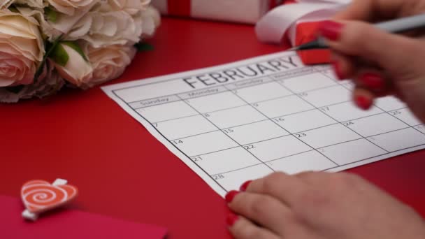 Las mujeres mano con marcador rojo pintan una forma de corazón en el calendario 14 de febrero. Vista lateral de una mesa roja con un ramo de rosas, una caja de regalo y un sobre festivo con un corazón. De cerca. Movimiento lento. — Vídeo de stock