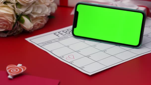 Widok z góry smartfona z zielonym klawiszem chroma na czerwonym stole w pozycji poziomej. Romantyczne tło z kalendarzem, w którym 14 lutego otoczony jest czerwonym sercem. Zamknij drzwi. Zwolniony ruch. — Wideo stockowe