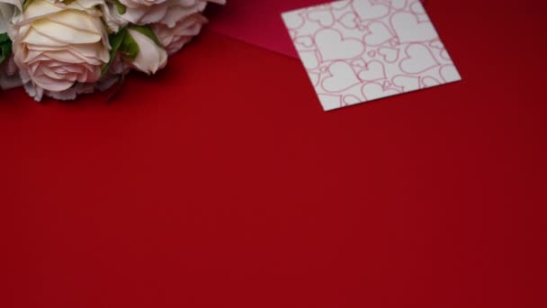 Gift box with a white ribbon bow falls on the red table. Festive background with attributes of Valentines Day, a bouquet of roses, an envelope, valentine and decorative hearts. Close up. Slow motion. — Stock Video