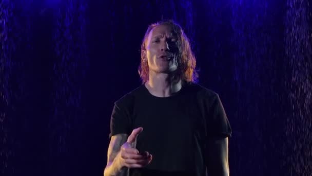 Músico de cabellos largos canta emocionalmente, gestos y se mueve expresivamente al ritmo de la música. Rocker actúa en vivo entre las gotas de lluvia en un estudio oscuro con luz azul. De cerca. Movimiento lento. — Vídeos de Stock