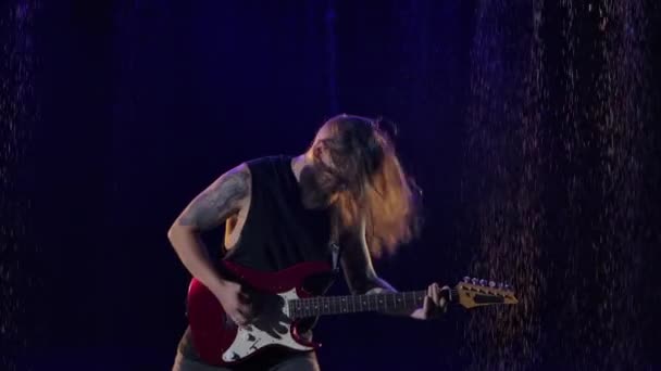 Musicista sta suonando parte solista sulla chitarra elettrica. Il musicista rock dai capelli lunghi suona magistralmente uno strumento a corda in uno studio buio sotto la pioggia. Concerto rock, musica dal vivo. Chiudete. Rallentatore. — Video Stock