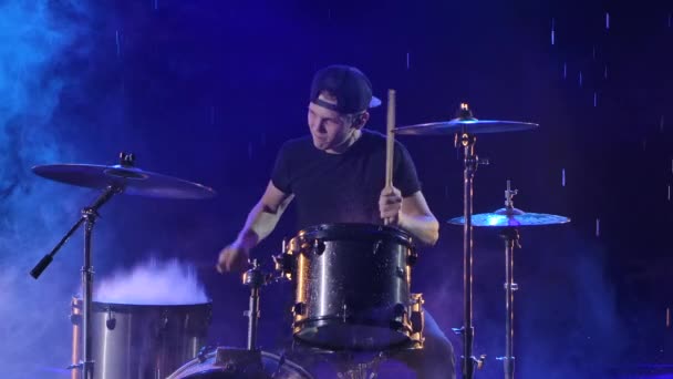 Young man enthusiastically plays the drum kit with drumsticks. A rock musician hits drums making music in the rain in a dark studio with blue lights. Close up. Slow motion. — Stock videók