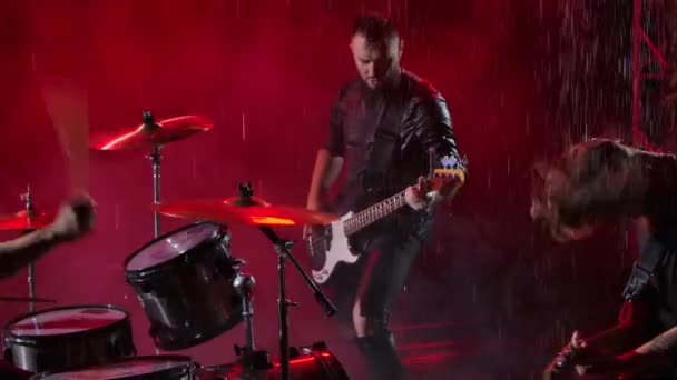 Camera glijdt over een groep rockmuzikanten die hun instrumenten met waanzinnige emoties bespelen in een donkere studio verlicht door rood licht. Drumstel, elektrische en basgitaar. Sluit maar af. Langzame beweging. — Stockvideo