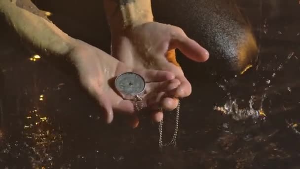 Man retrieves a drowned compass from the water. Wet person kneels on surface of the water in pouring rain. Shot in a dark studio illuminated by yellow lights. Close up. Slow motion. — Stock video