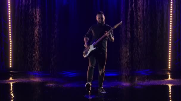 Le musicien barbu de rock joue magistralement en solo sur la guitare basse dans un studio sombre avec des lumières bleues. Le guitariste se produit en direct sous la pluie battante. Mouvement lent. — Video