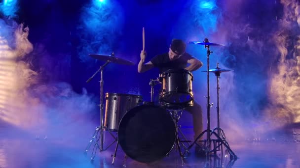 Jovem joga entusiasticamente o kit de bateria com baquetas. Um músico de rock toca bateria fazendo música à chuva em um estúdio escuro fumegante com luzes azuis. Movimento lento. — Vídeo de Stock