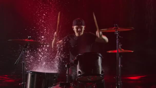 Progresivní mladý muž nadšeně hraje na bicí soupravu s paličkami. Rockový muzikant bubnuje v dešti v zakouřeném tmavém studiu s červenými světly. Šplouchání a cákání vody stoupá zpomaleným pohybem vzhůru. — Stock video