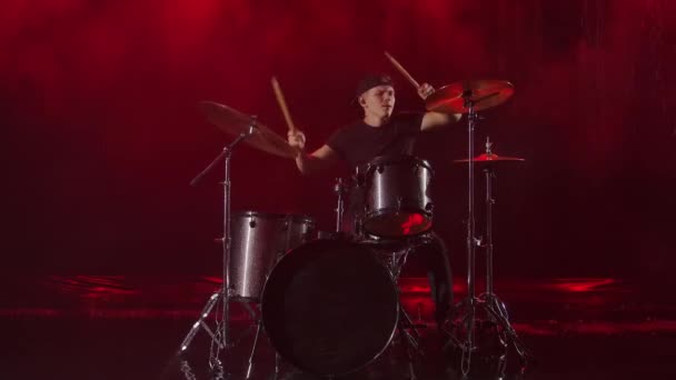 Jongeman speelt enthousiast de drumkit met drumsticks. Een rockmuzikant slaat drums die muziek maken in de regen in een rokerige donkere studio met rood licht. Langzame beweging. — Stockvideo