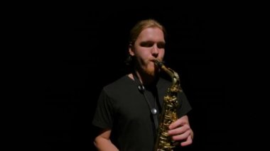 Kameralar saksafon çalan uzun saçlı genç adamın etrafında ağır çekimde dönüyor. Müzisyen karanlıkta rüzgar enstrümanı çalmayı sever. Stüdyo ışıklarıyla siyah arka planda izole edilmiş. Kapat..