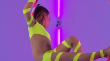 Genç kız hava akrobatları havada bir çemberin üzerinde döner. Stüdyodaki parlak neon ışıkların arka planına karşı sarı taytlı bir sirk sanatçısı. Kapatın. Yavaş çekim.