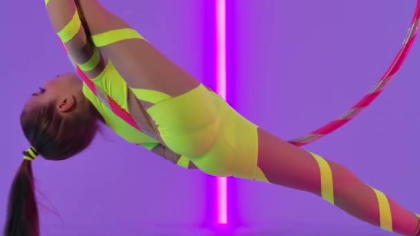 Young aerial acrobat girl rotates in the air on a hoop. Circus performer in a yellow leotard against the background of bright neon lights in the studio. Close up. Slow motion. — Stock Video