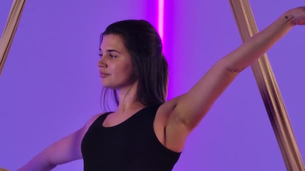 La gimnasta aérea femenina flexible realiza rotaciones y divisiones en la seda aérea. Una joven en un maillot negro se equilibra contra el fondo de brillantes luces de neón en el estudio. De cerca. Movimiento lento. — Vídeo de stock