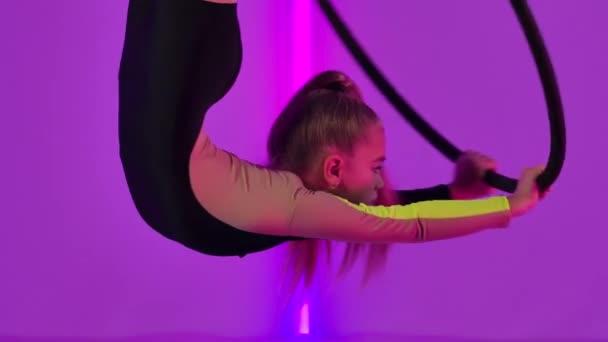 La gimnasta joven y flexible cuelga boca abajo en un aro de aire y realiza elementos acrobáticos. Chica en maillot negro realiza espectáculo de circo contra el telón de fondo de luces de neón brillantes. De cerca. Movimiento lento. — Vídeos de Stock