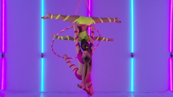 Dos chicas jóvenes realizan elementos acrobáticos en un aro de aire. Acróbatas aéreas en un espectáculo de circo en el telón de fondo de luces de neón brillantes. Movimiento lento. — Vídeos de Stock
